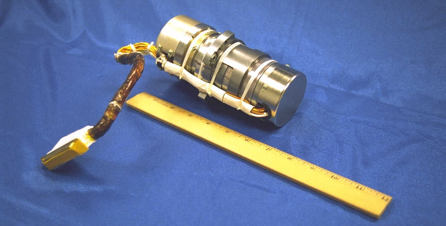 A Hubble gyro in its metal container with a bundle of electronic wires and a connector extending outside the cylinder. The unit sits next to a yellow ruler to show it is approximately one half foot long. All are on a blue background.