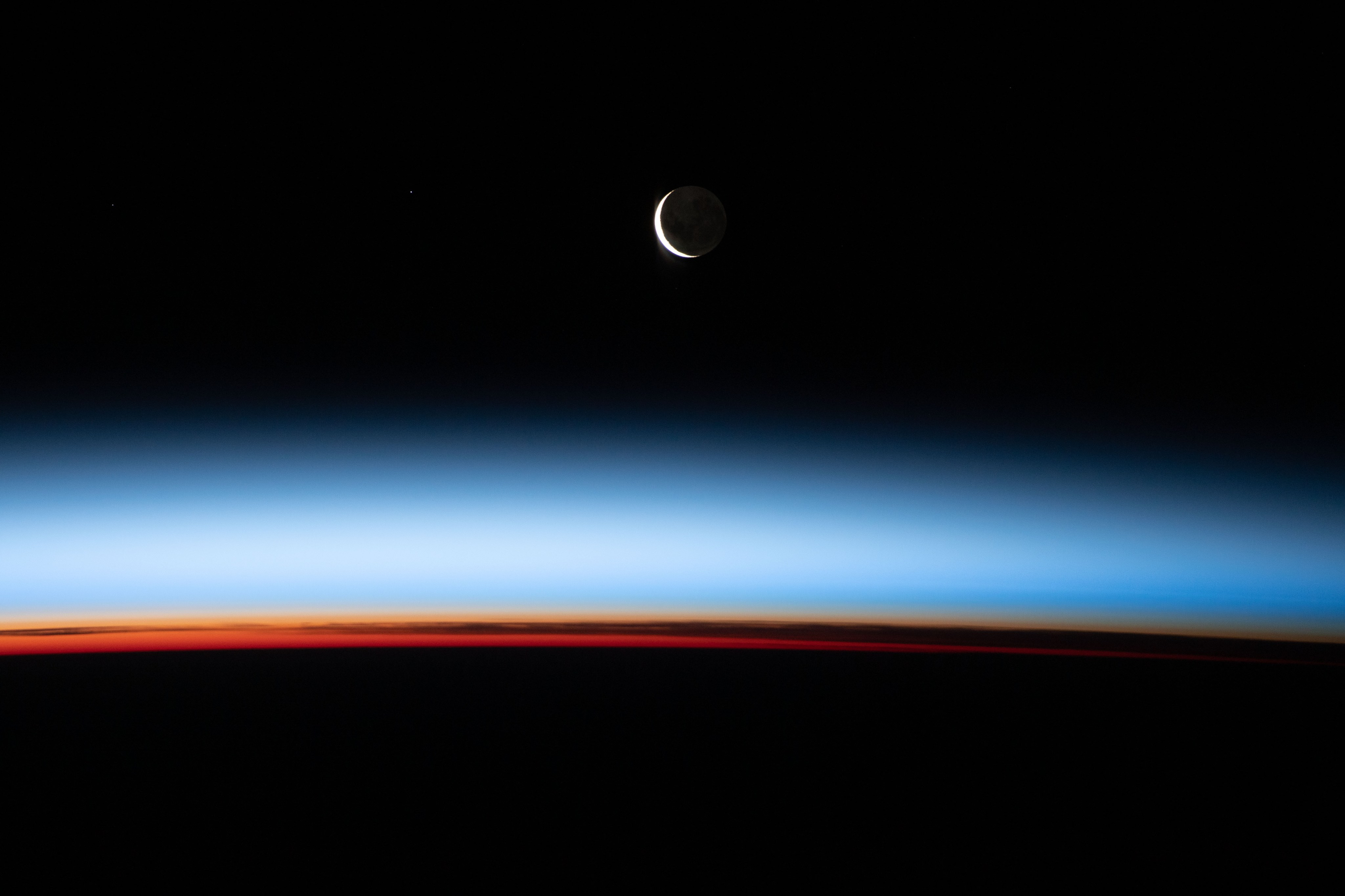 Photograph of a crescent moon in space. Earth's horizon, topped by a thin, glowing layer of atmosphere, fills the bottom edge of the picture.