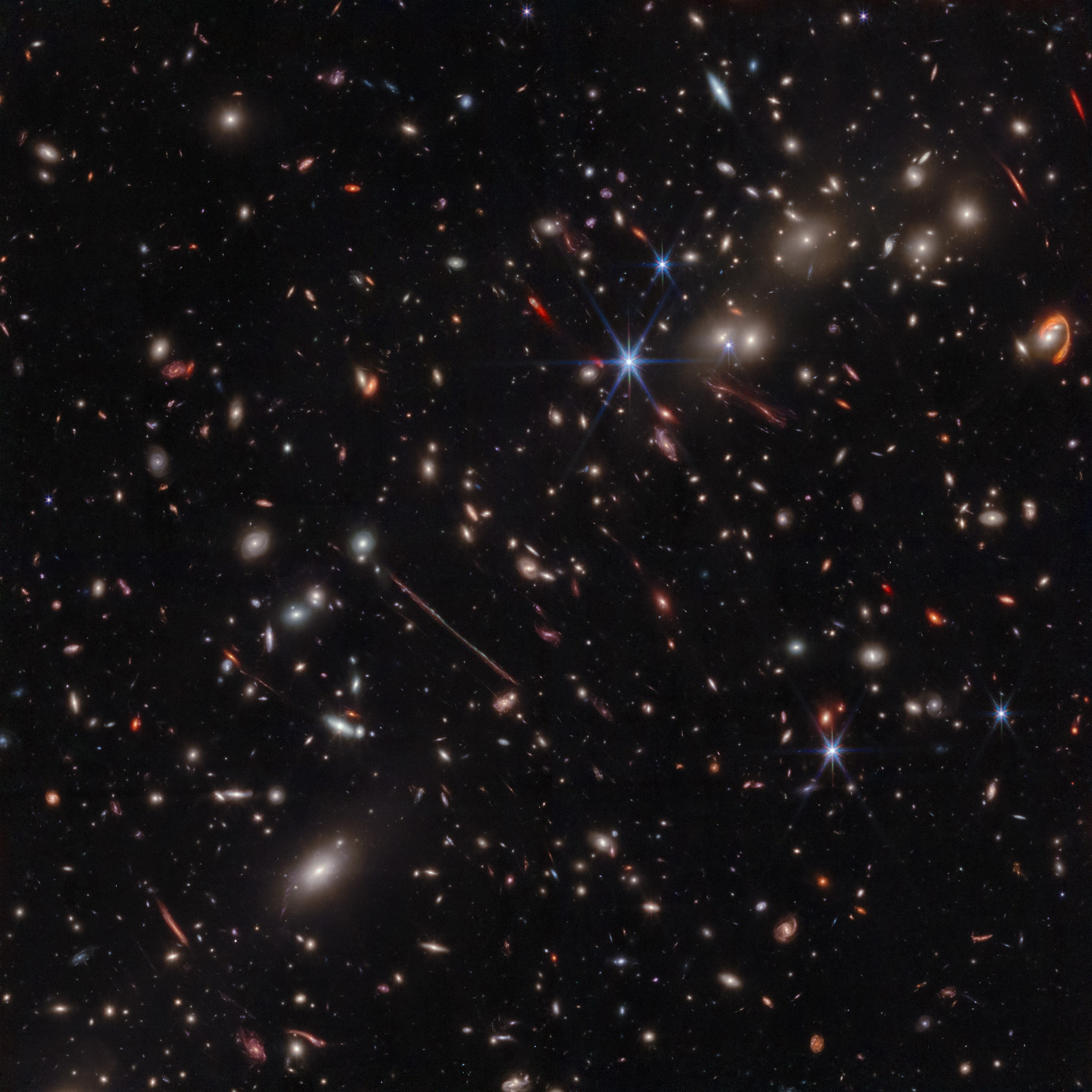 A black background is scattered with hundreds of small galaxies of different shapes, ranging in color from white to yellow to red. Some galaxies are distorted, appearing to be stretched out or mirror imaged. Near the center, a particularly long and thin line stretches from ten o’clock to four o’clock. At upper right, a red swoosh extends about three-quarters of the way around a pair of galaxies. A handful of foreground stars display eight diffraction spikes.