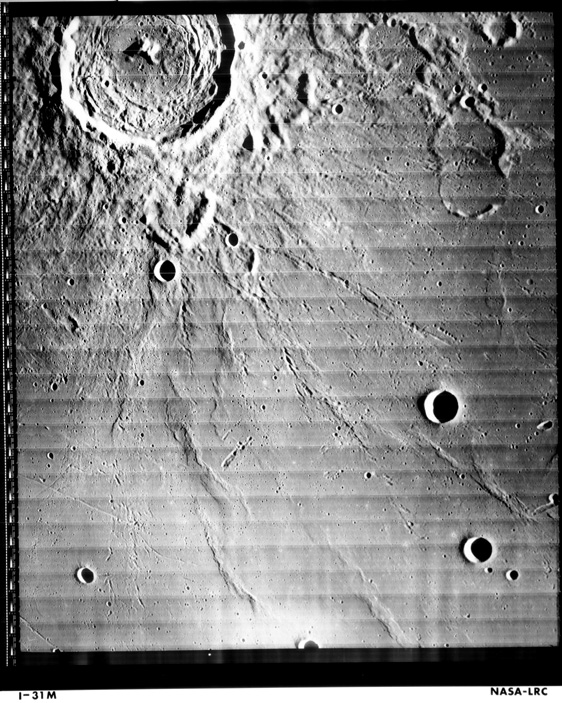 Off-centered photo of clearly- defined crater on the Moon, with ridges and smaller craters extending from the large crater outwards. 