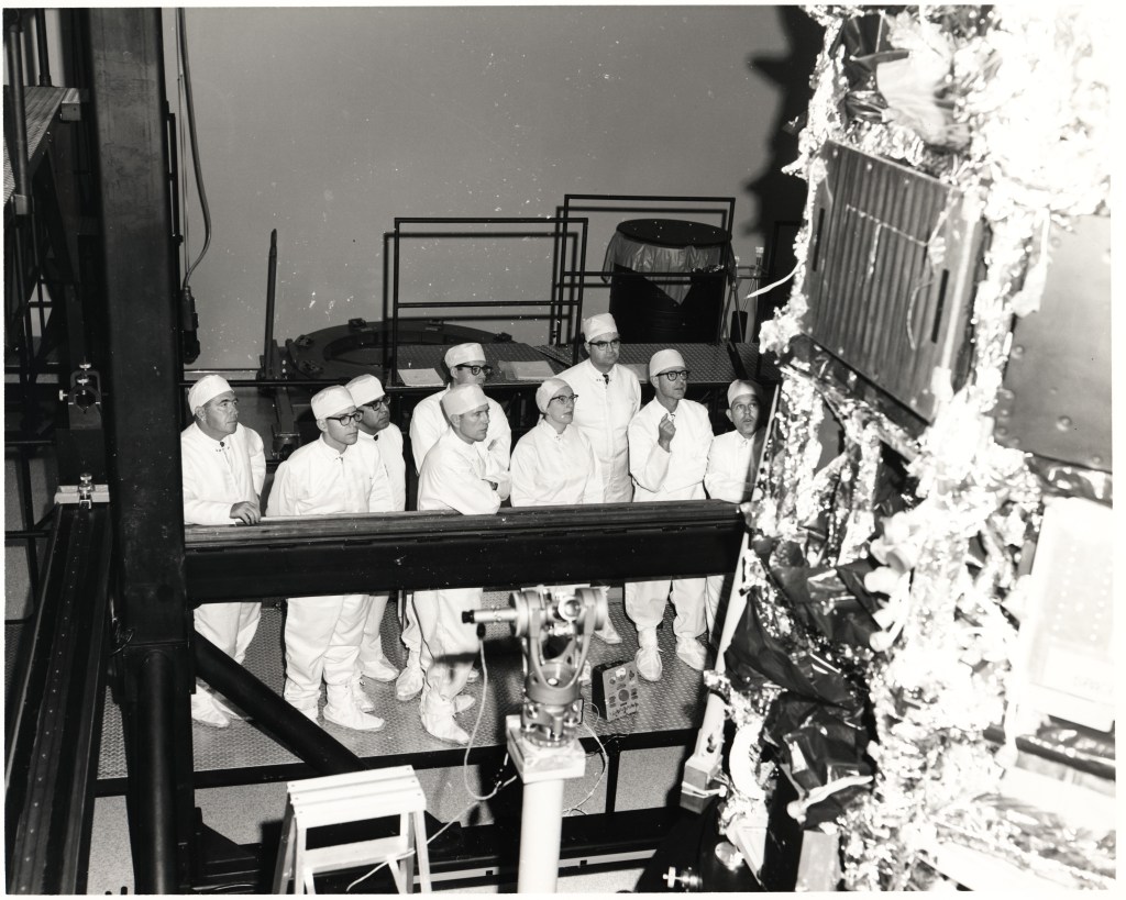 A group of people in clean room suits stand next to a spacecraft in 1964.