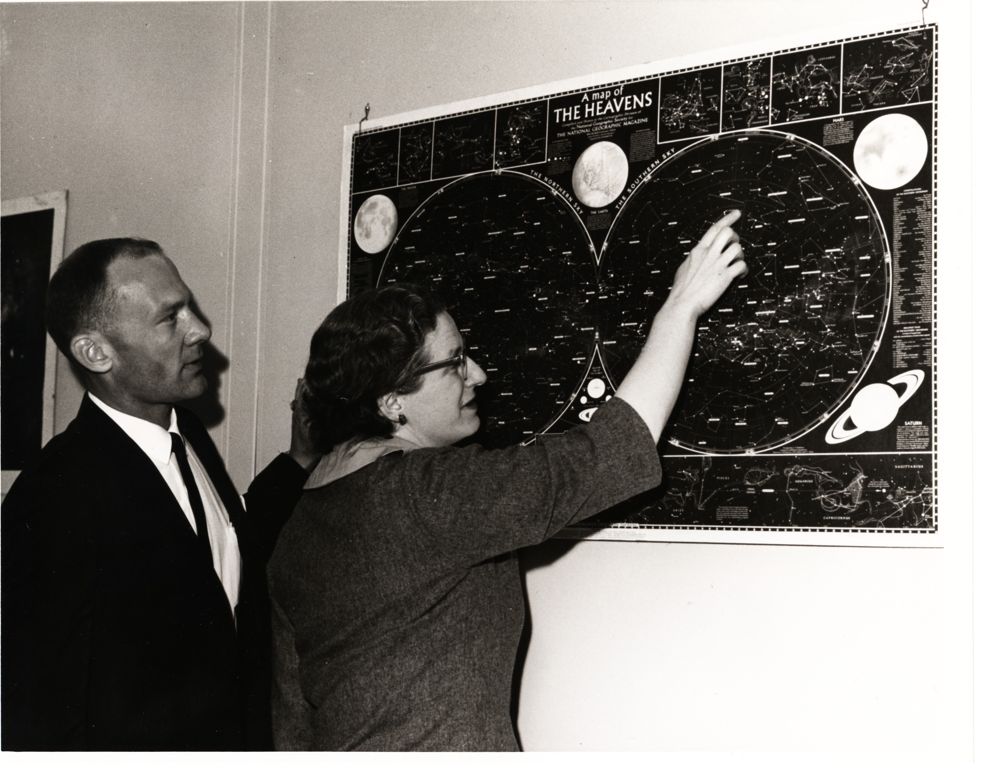 Nancy Grace Roman points on a sky map that is on a wall while Buzz Aldrin looks on.
