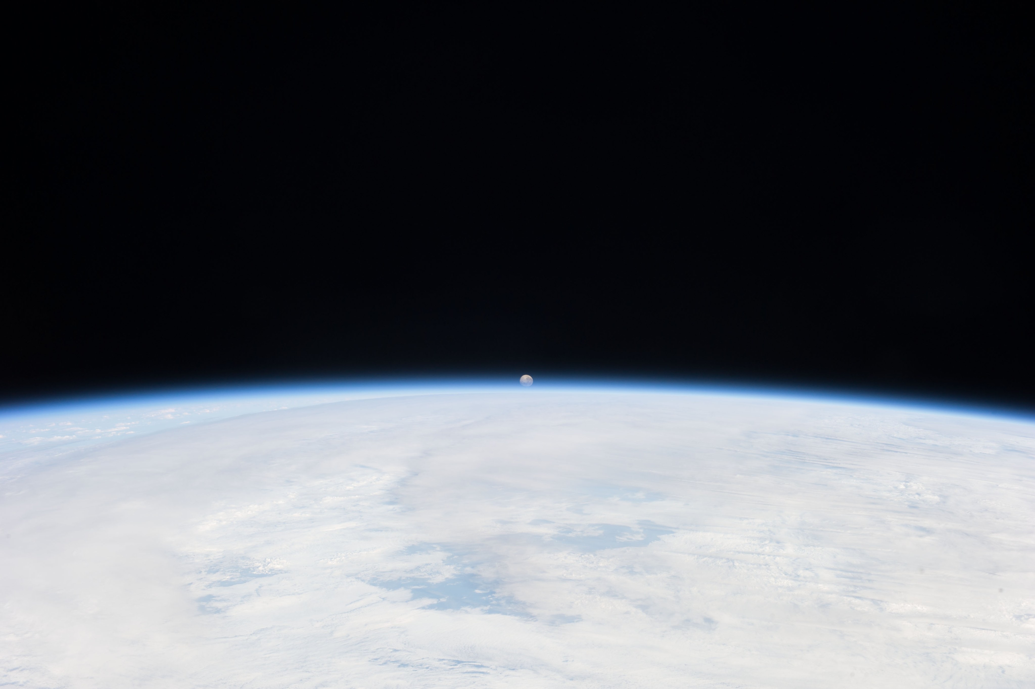 Tiny-looking Moon setting in the distance over Earth's curving, cloud-covered horizon.