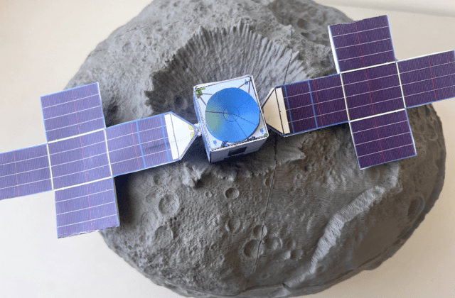 A paper model of the Psyche spacecraft with its solar wings unfurled. An illustration of the asteroid is behind the spacecraft model.