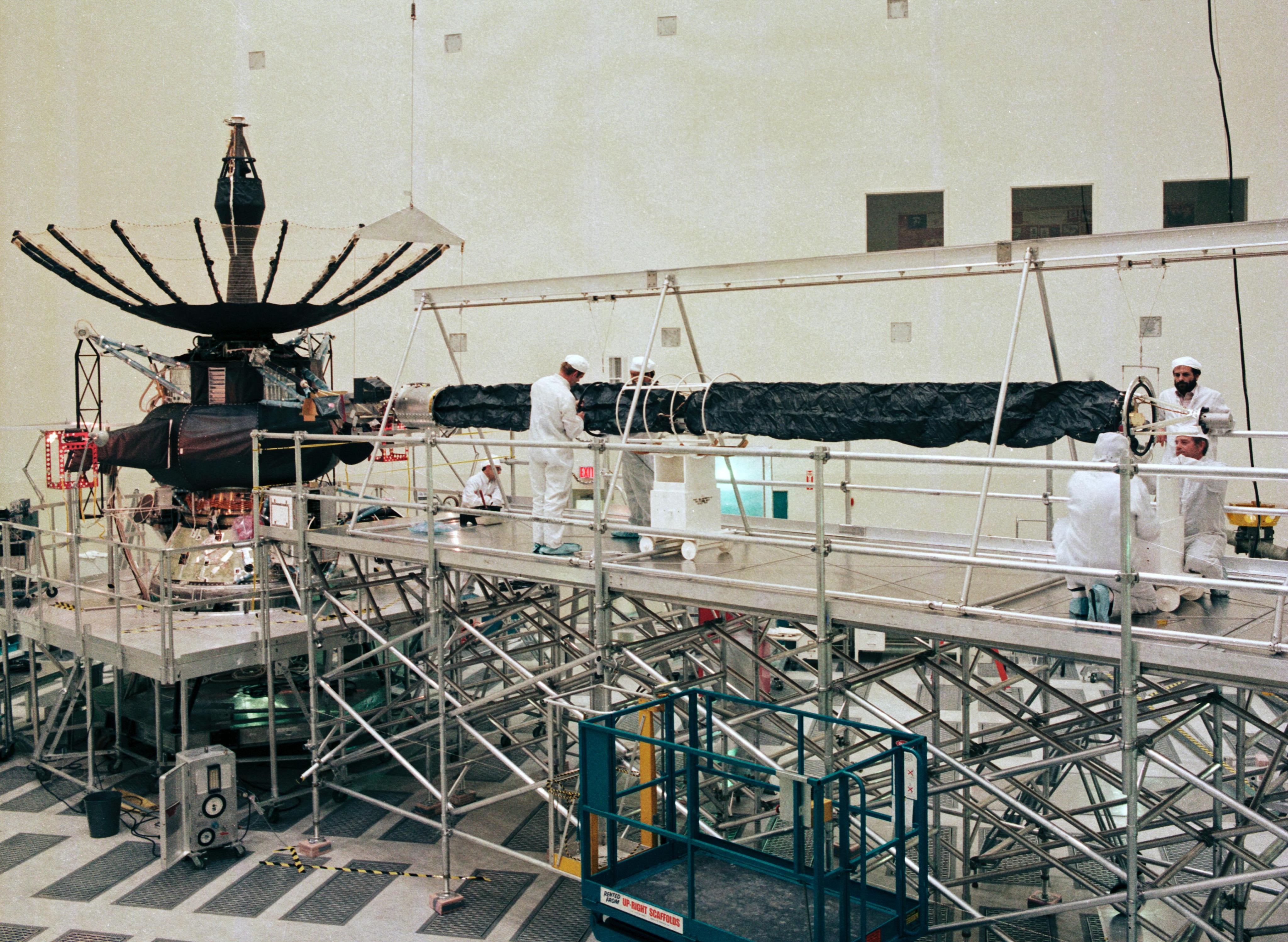 Engineers in white cleanroom suits stand on scaffolding to work on the fully-assembled Galileo orbiter.