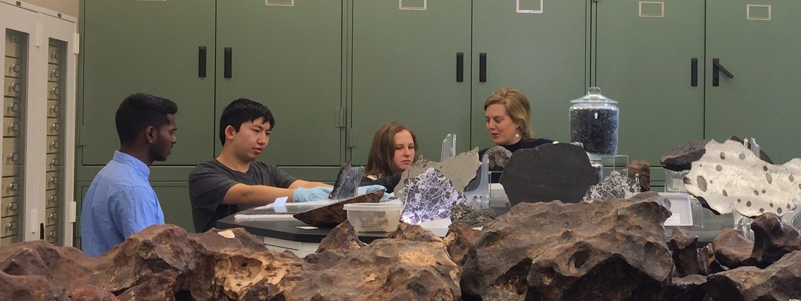 Four students stand behind a table covered with rocks and other materials for projects.