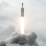 The Psyche spacecraft rides to space inside its faring on top of a rocket with two side boosters. The spacecraft faring and boosters are all white. Flames shoot out the bottom of the rocket stack, and clouds are beneath the flames.