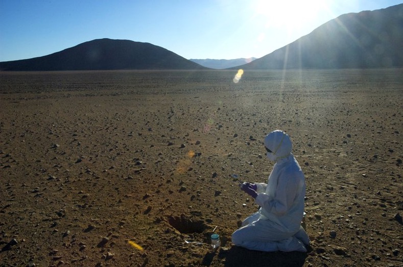 At the center of this image is a scientist wearing a white clean suit and mask, as well as gloves and goggles. There is a small hole and sterile tools in front of the scientist, who kneels in the soil of the Atacama desert. A desert landscape similar to that of Mars surrounds the scientist, with a vast stretch of dry, rocky soil leading up to shadowed mountains. Above the mountains and set amid a blue sky, the Sun peeks out at the top right of the image, causing a lens flare that stretches across the image.
