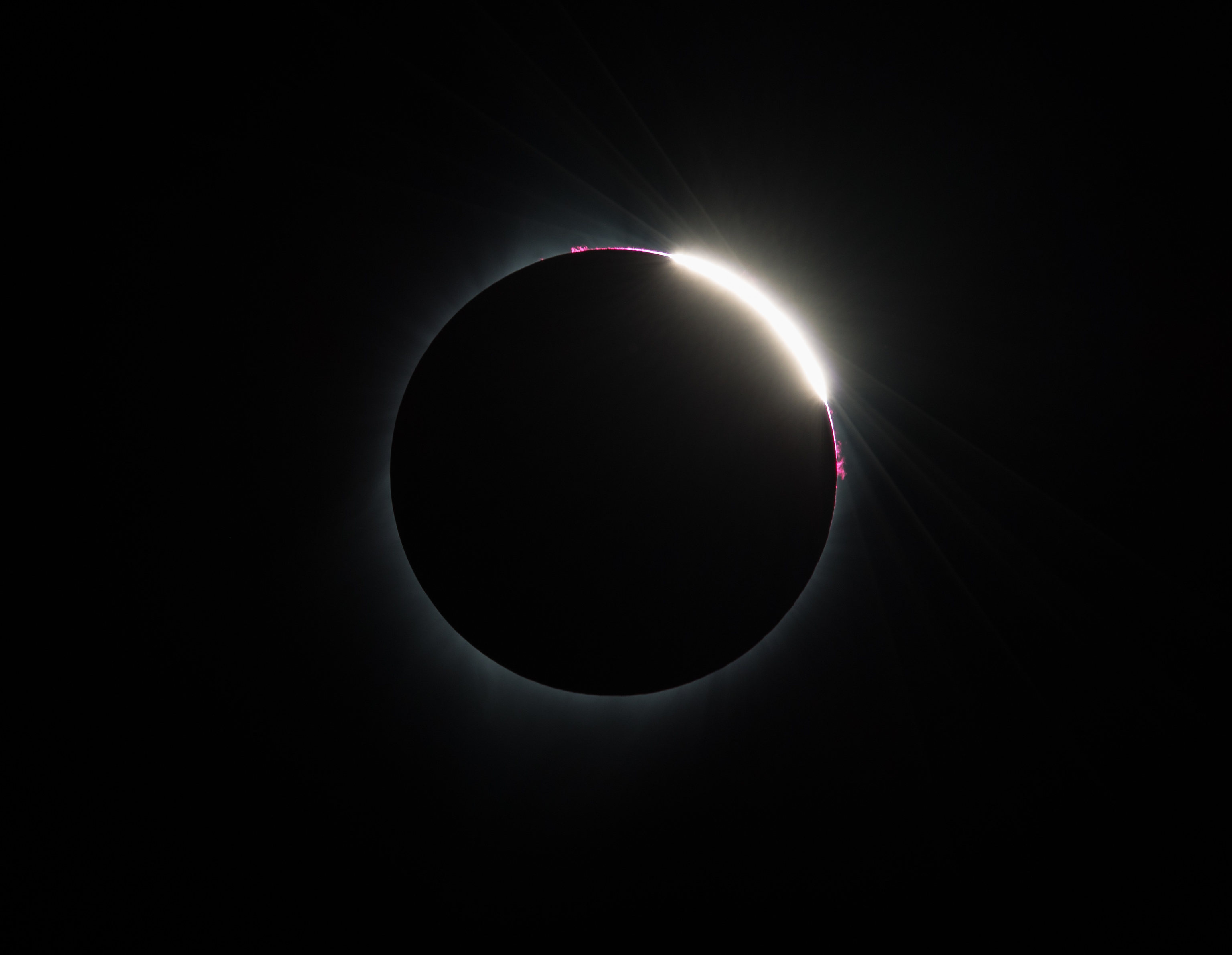 Against a black background is a solar eclipse. There is a large black circle in the middle of the image – the Sun. On the top right of the circle is a bright white sliver of light from the Sun. On either side of the sliver of light are small spots of pink looping away from the circle – prominences.