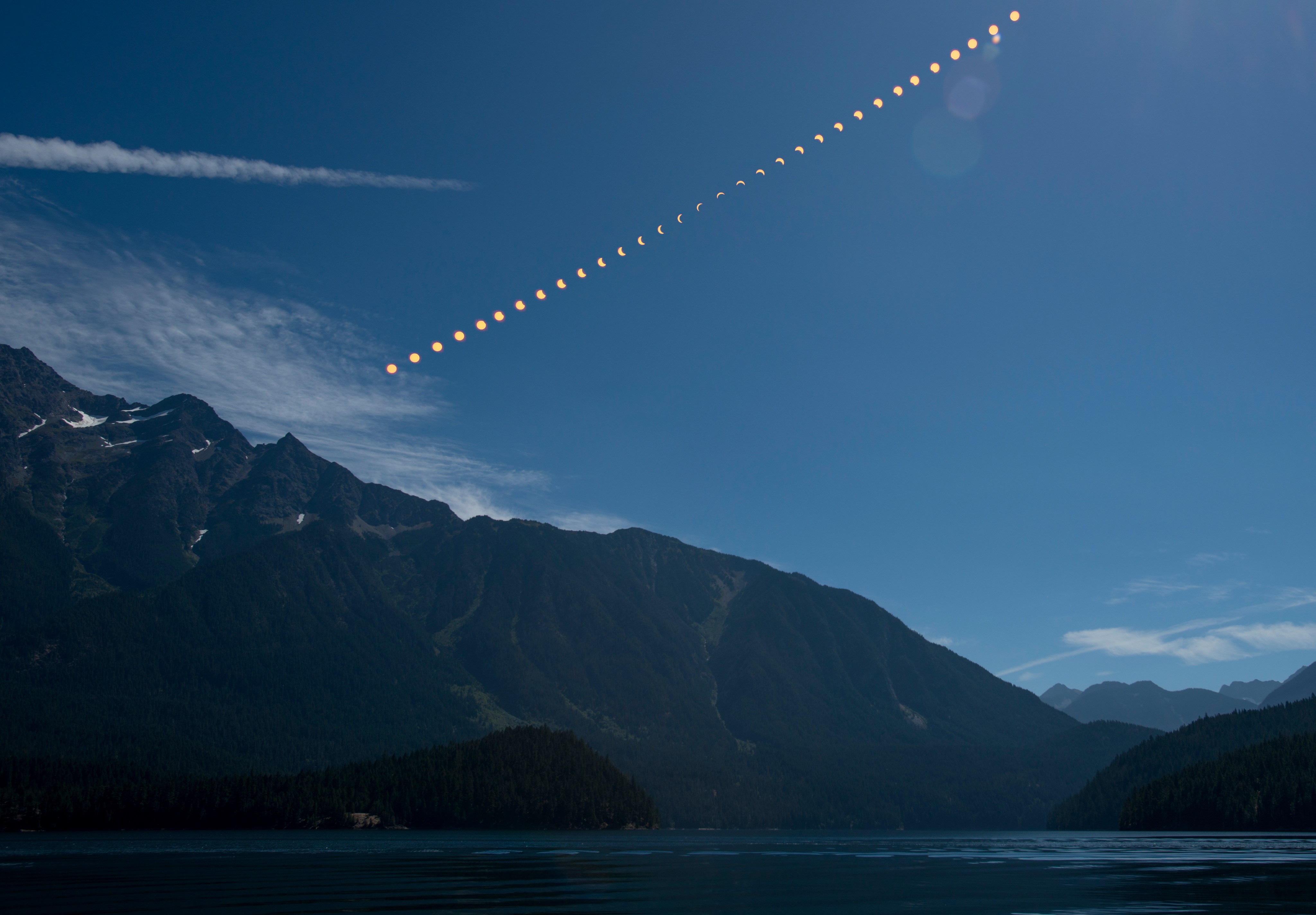 A composite image showing mountains above a lake and blue sky. In the sky is a total eclipse, progressing from a full Sun, to totality, and back to a full Sun again.
