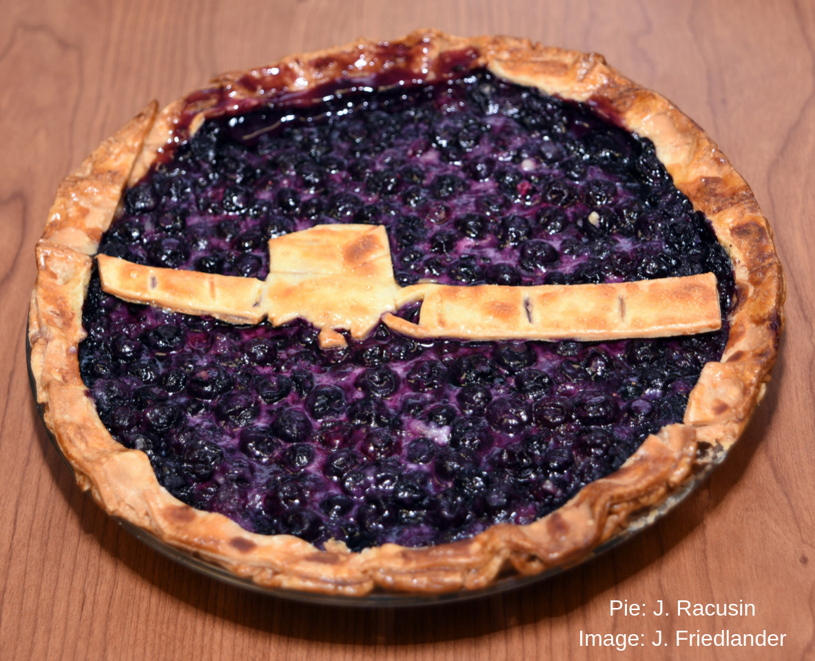 This image shows a pie decorated to celebrate the Fermi Gamma-ray Space Telescope sitting on a wood-grained brown background. The pie is in a silver pie plate with a tan crimped outer crust and is filled with shiny purplish blueberries. A simple two-dimensional model of Fermi made of pie crust spans the center of the pie. It is shaped like a box with long solar panels extending from it, with scored lines to add details. The image is watermarked with the text “Pie: J. Racusin” and “Image: J. Friedlander.”