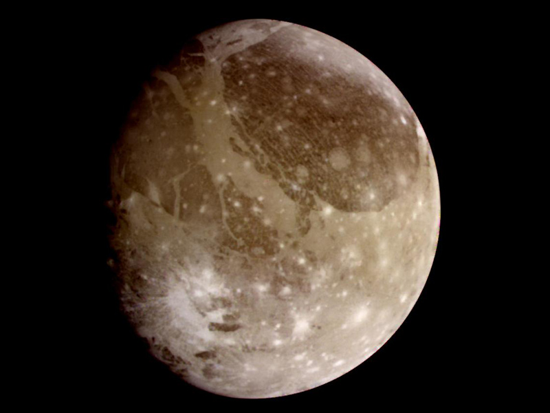 A brownsh gray sphere, shaded on the left side, appears against a black background. It's mottled with dark patches in the upper half, while the lower half is spotted with beight, white mars, some with rays extending from them like paint splatters.