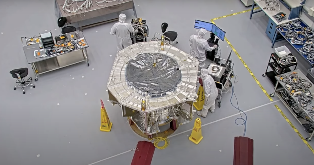 Three people in all white suits, which cover their entire body, stand around the IMAP spacecraft on the floor. 