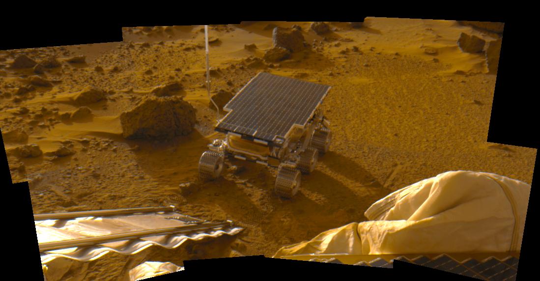 In the middle of the frame a small, six wheeled rover, with its entire top consisting of a solar panel, sits on terrain of rust-colored dirt and rocks. The silver ramp it just descended is visible in the lower left.