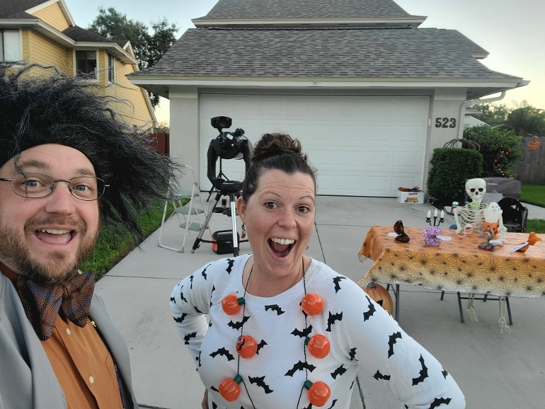 two people do a happy face while dressed up in Halloween garb in front of a telescope and table with a telescope