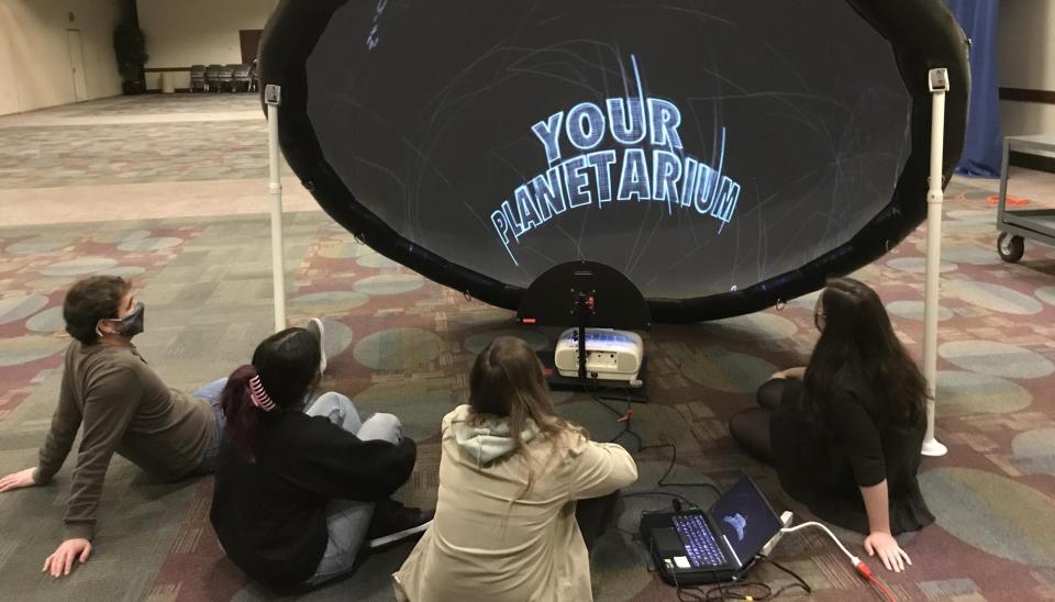 Photo of kids sitting on the floor in front of a large round screen that reads "Your Planetarium"