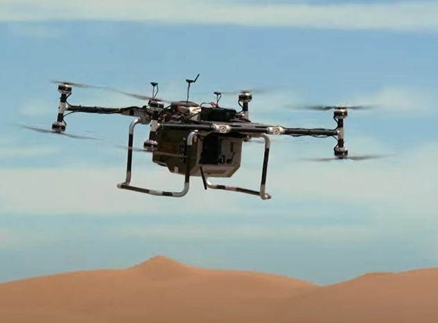 A quadcopter is seen flying above brown sandy dunes with a partly cloudy sky in the background.