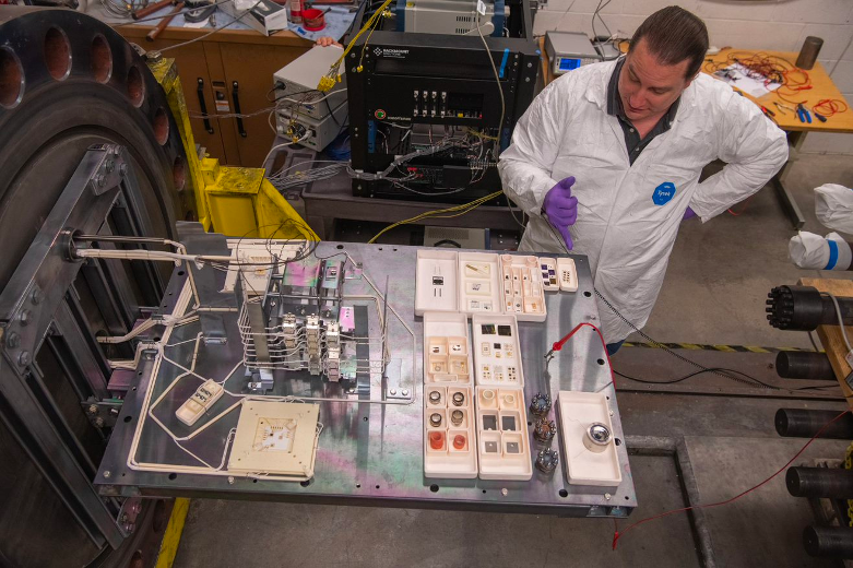 A person in a white lab coat and gloves standing next to rectangular platform that holds the test articles and is attached to the test chamber.