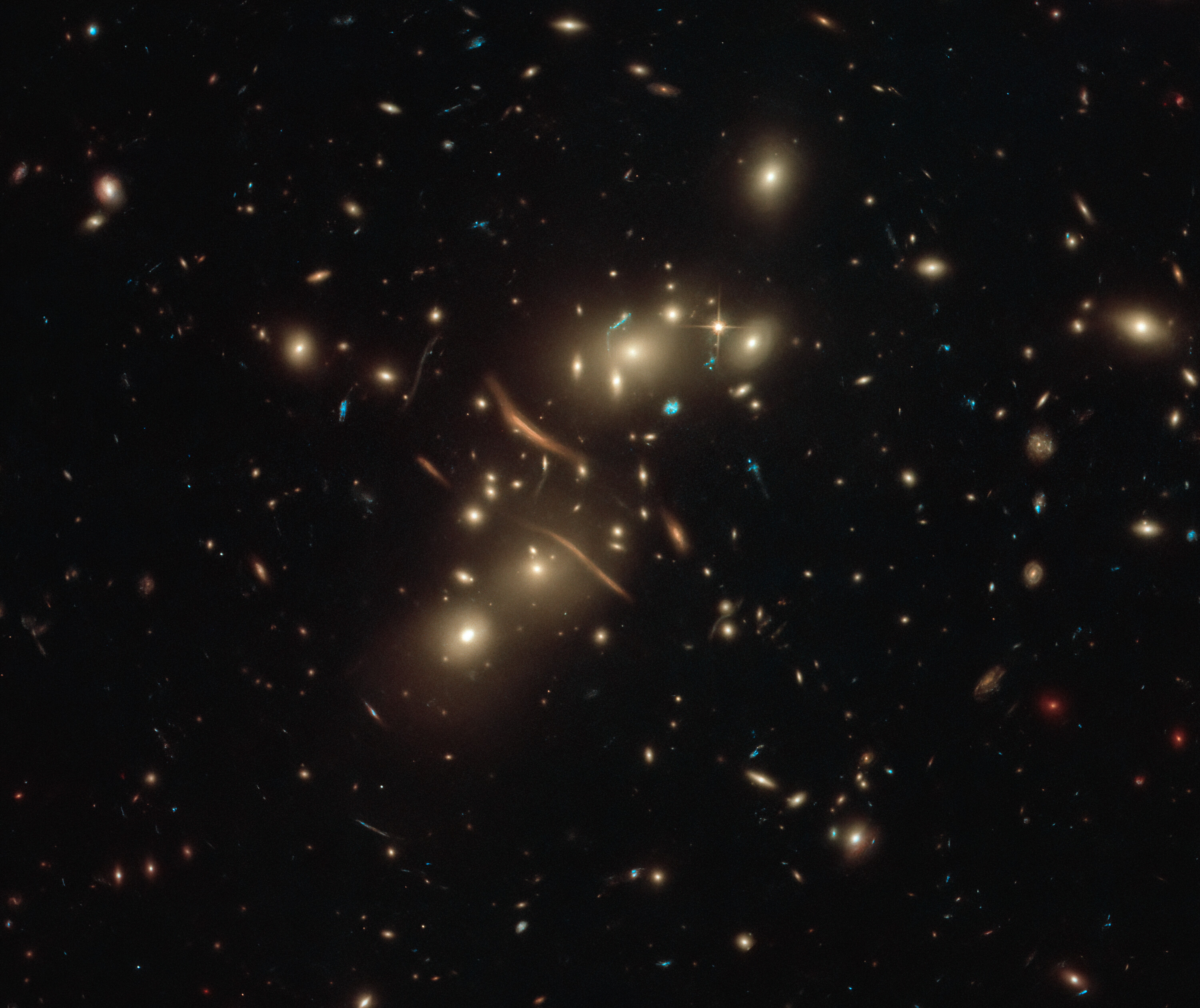 Black background is dotted with distant galaxies. Image center holds a cluster of galaxies, many of the appear to be large elliptical galaxies. Curved orange-yellow lines appear to cut through the galaxy cluster. These are more distant galaxies gravitationally lensed by the cluster.