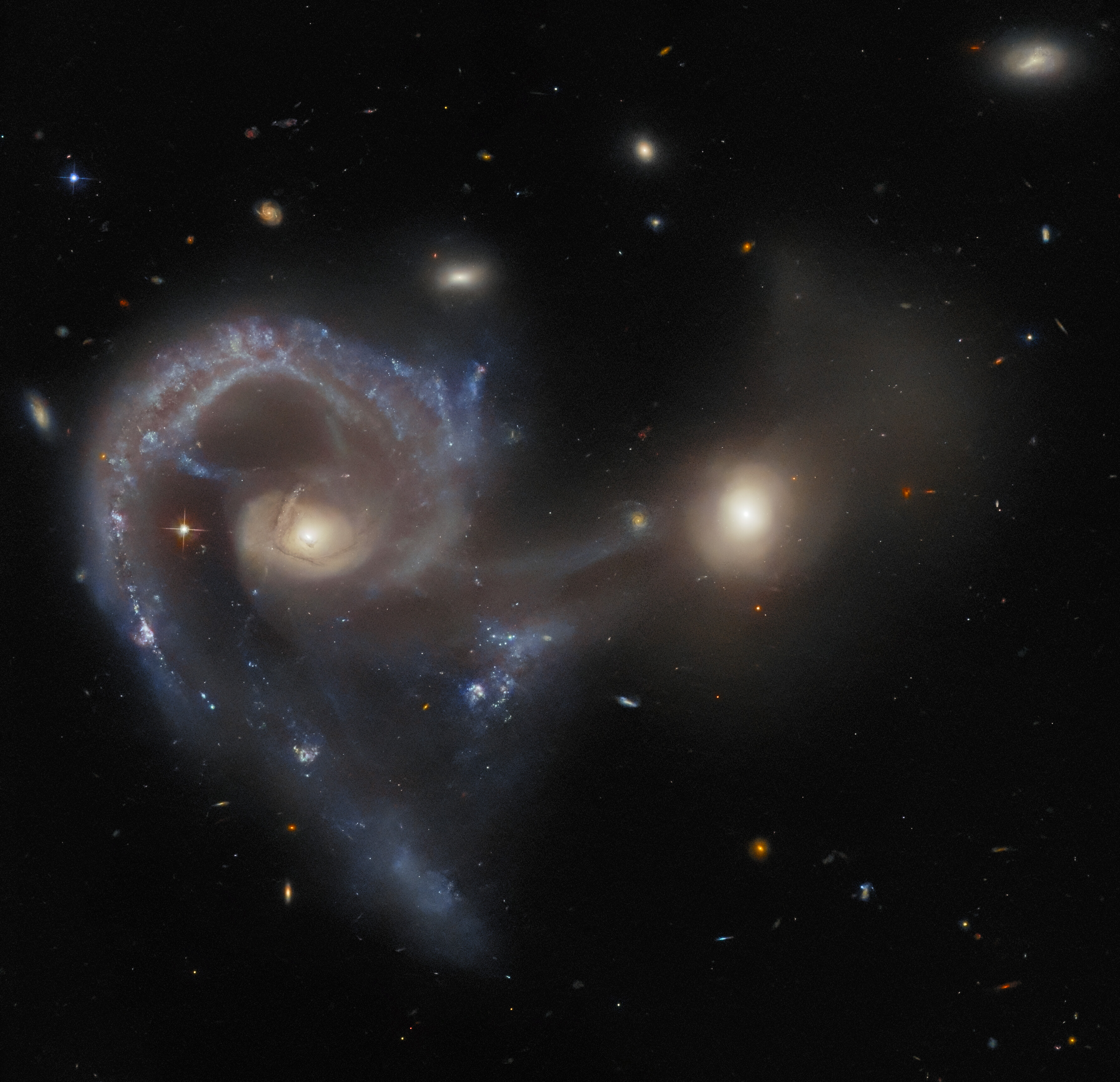 A pair of merging galaxies. The galaxy on the left has a large, single spiral arm curving out from the core and around to below it, with very visible glowing dust and gas. The right galaxy has a bright core but only a bit of very faint material. A broad curtain of gas connects the two galaxies’ cores and hangs beneath them. A few small stars and galaxies are scattered around the black background.