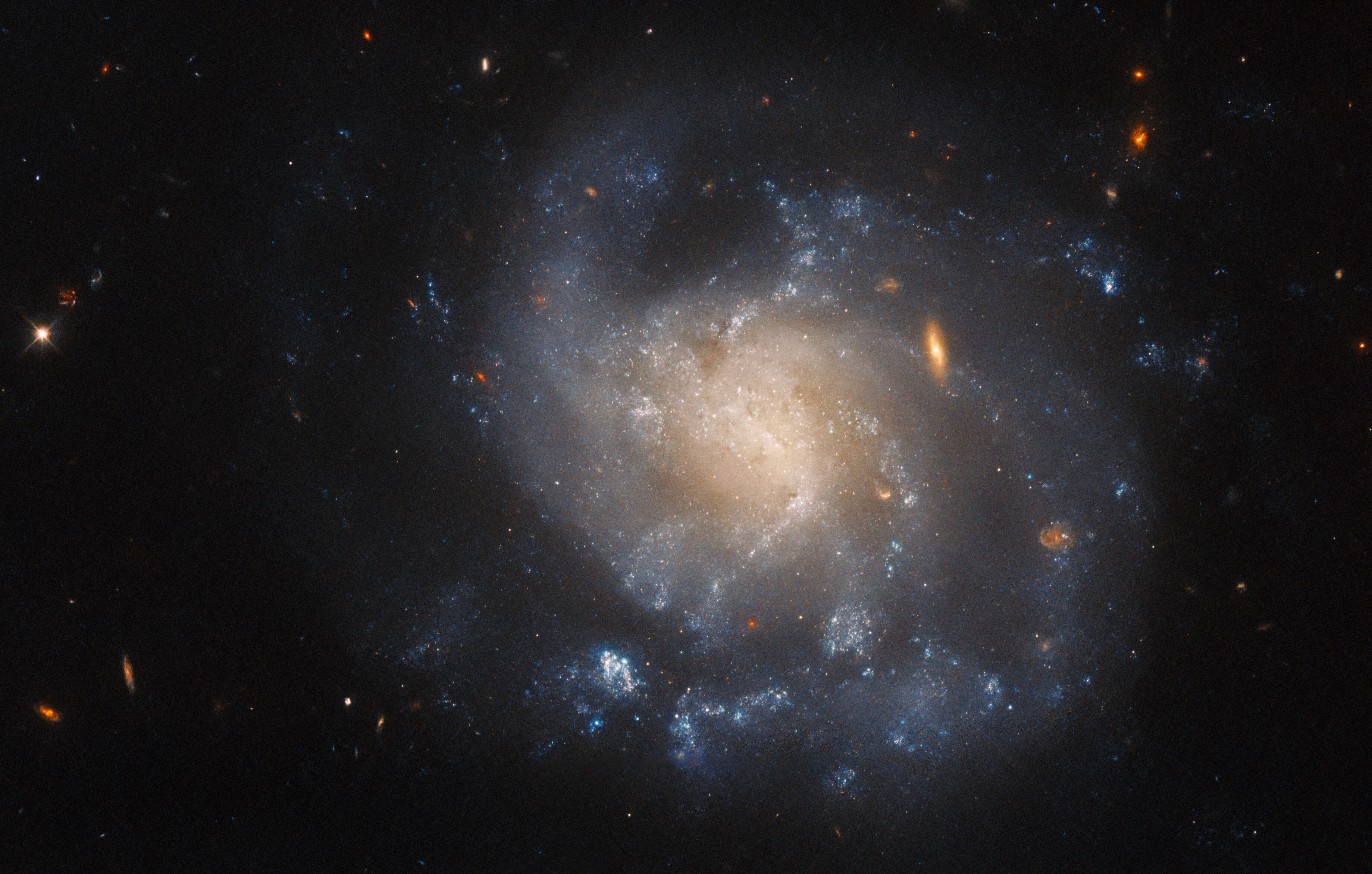 An irregularly-shaped spiral galaxy. Its spiral arms are difficult to distinguish. The edges are faint, and the core has a pale-yellow glow. It is dotted with small, wispy, blue regions where stars are forming. A few stars and small galaxies in warm colors are visible around it.