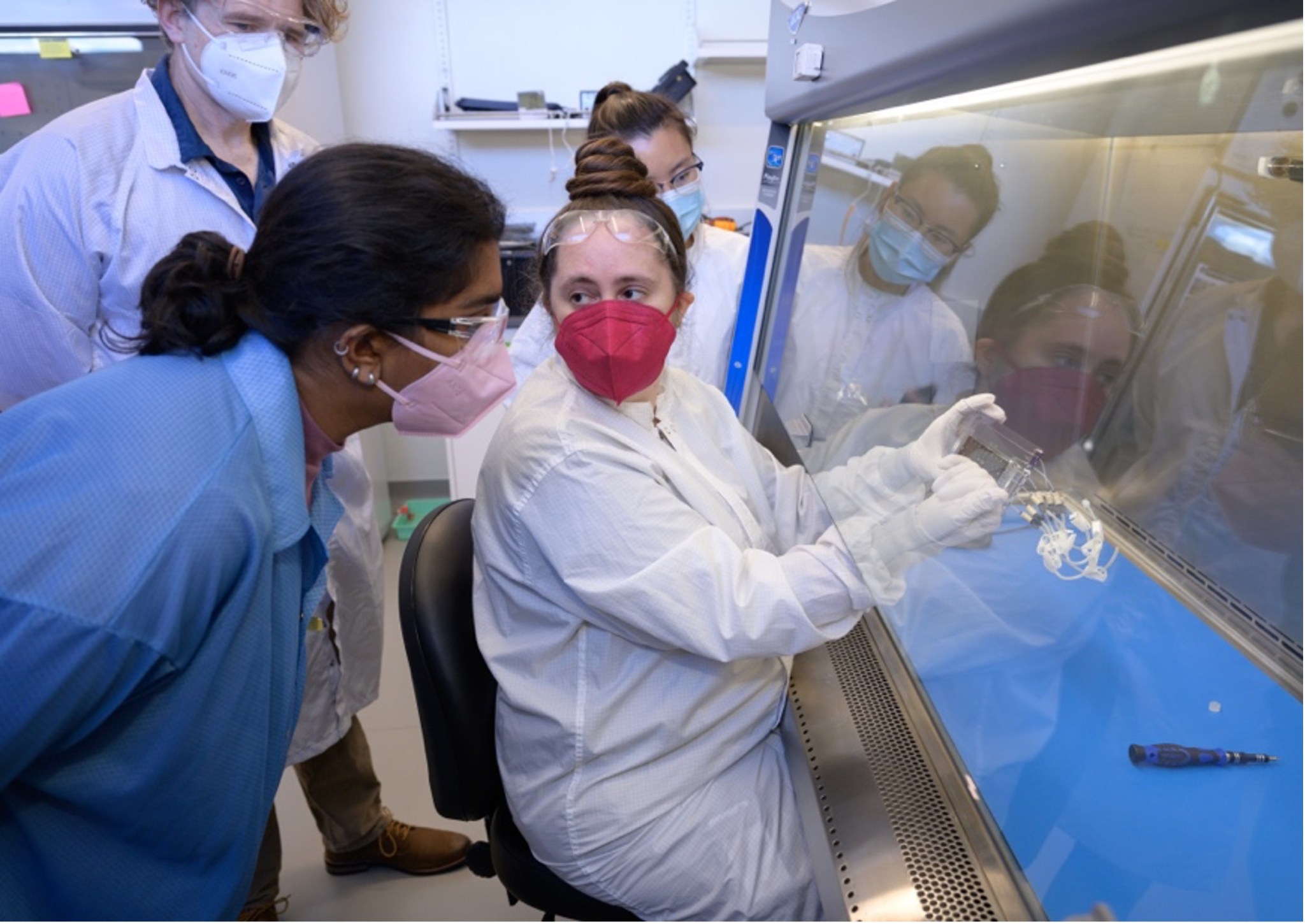 Three team members wearing lab coats and face masks; one team member is seated with gloved hands above a special work table, and the other two are watching.