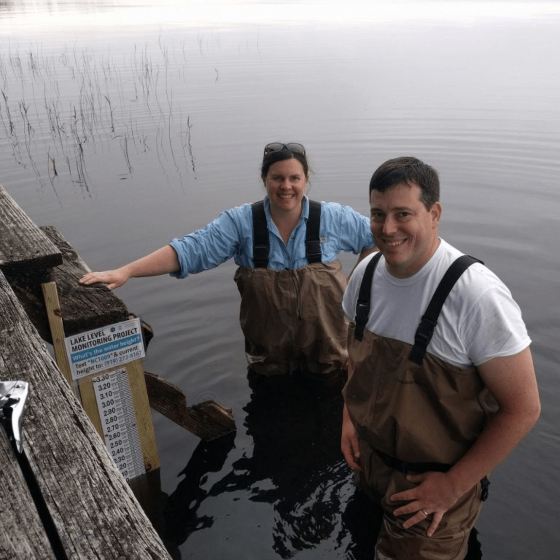 A man and a woman, each wearing brown overall waders and smiles, are standing in thigh-high water next to a dock. On the dock, they have recently installed a water level gauge, which looks like an oversized ruler extending above and below the surface of the lake. 