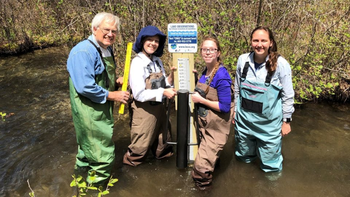 A grey-haired man and three young women, all in waders adn all with big smiles, stand knee-deep in water next to a LOCSS lake level gauge they are in the process of installing. The gauge itself is partially blocked from view by a black post, presumably the one on which they will mount the new gauge.