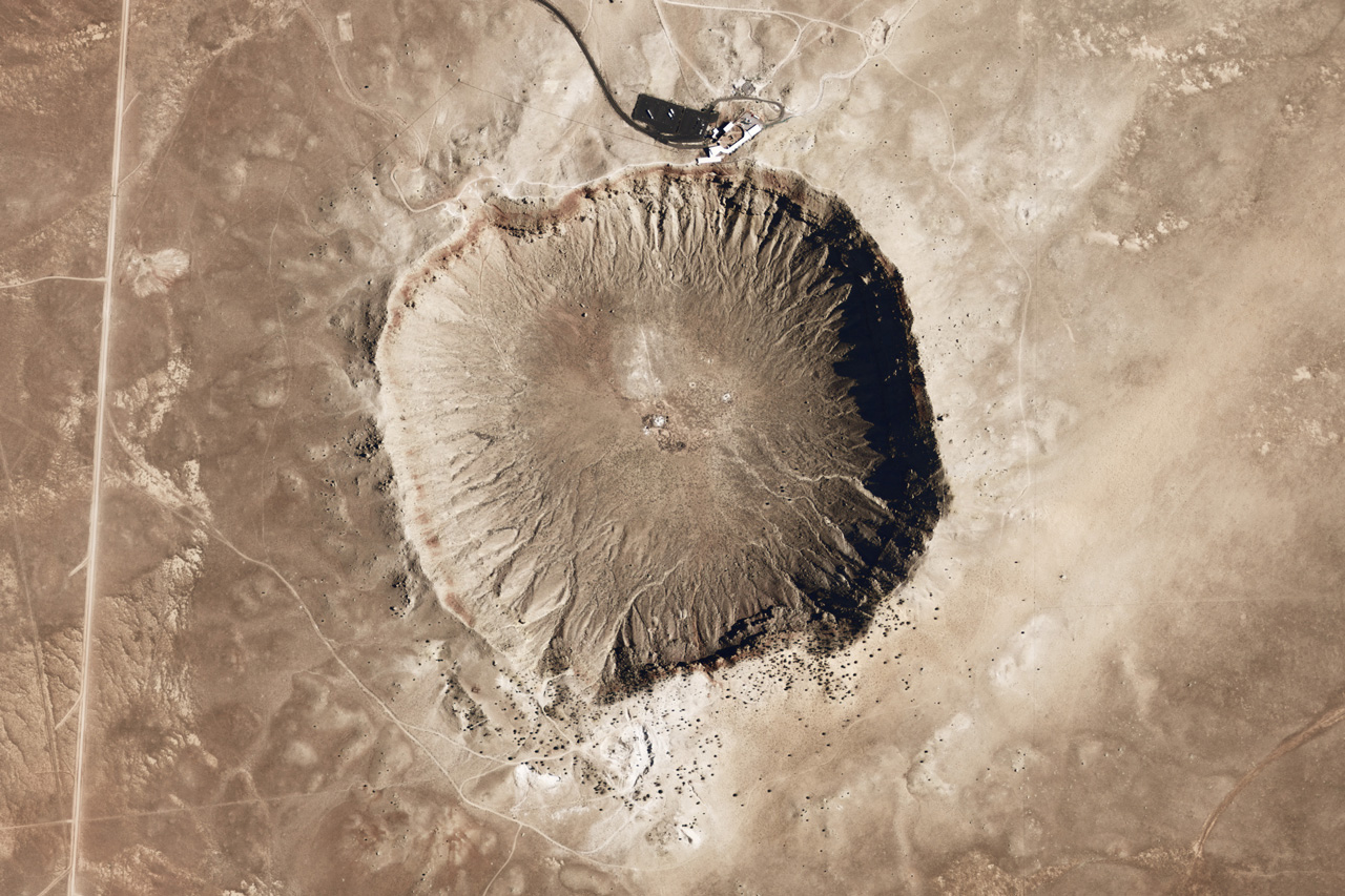 Giant crater with brown earth around it. The visitor's center and cars are visible at the top of the image.