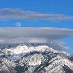 A bright full moon in a blue daytime sky over snow-capped peaks.