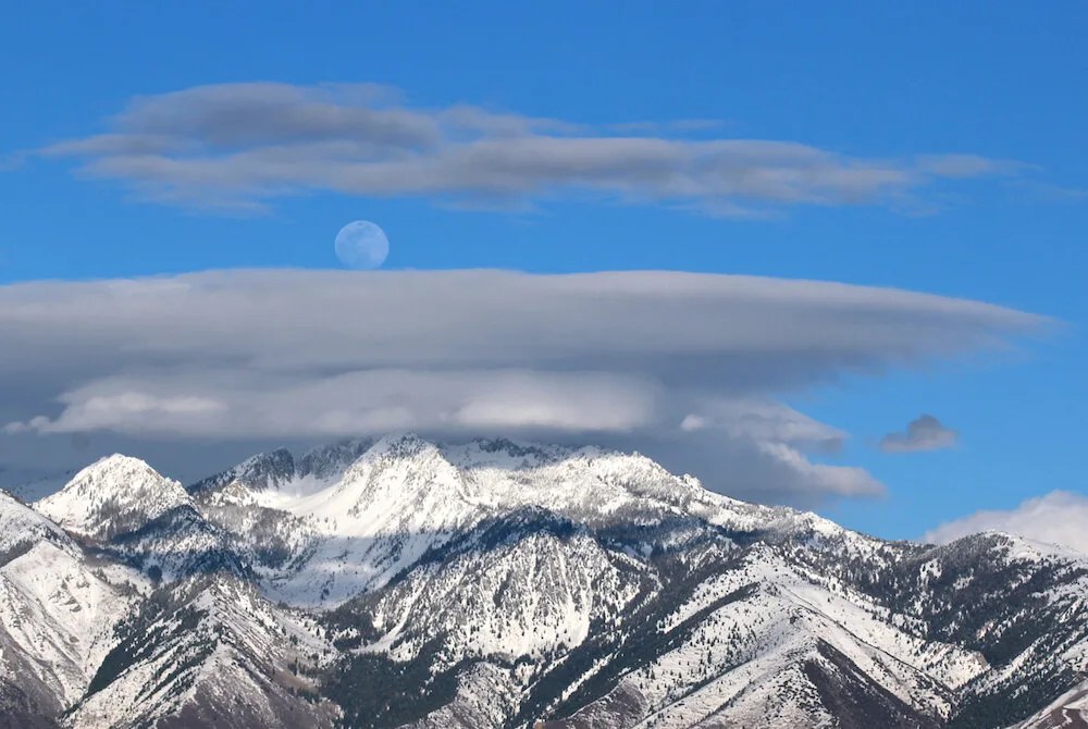 A bright full moon in a blue daytime sky over snow-capped peaks.