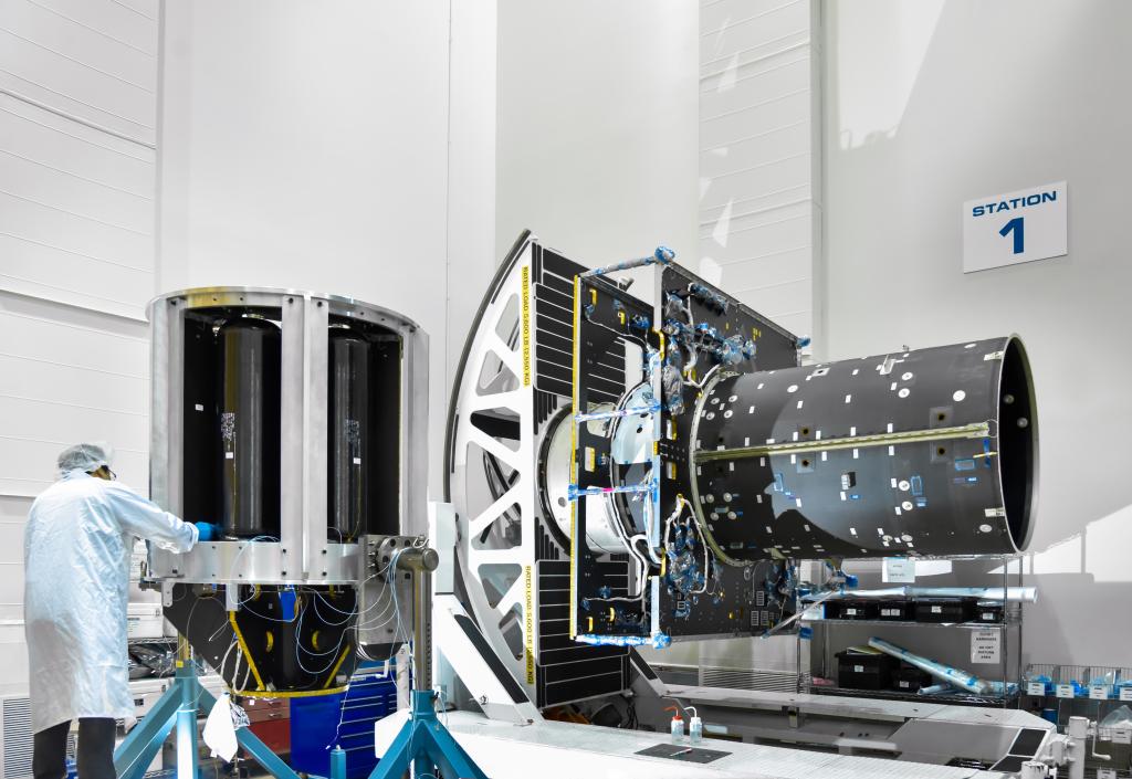 A worker with their back to the camera works on the Psyche spacecraft. Two large pieces of the spacecraft sit on the floor in front of the worker.