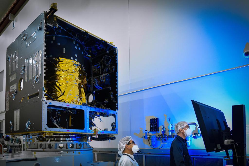 Engineers in protective clothing work near the Psyche spacecraft's main body.