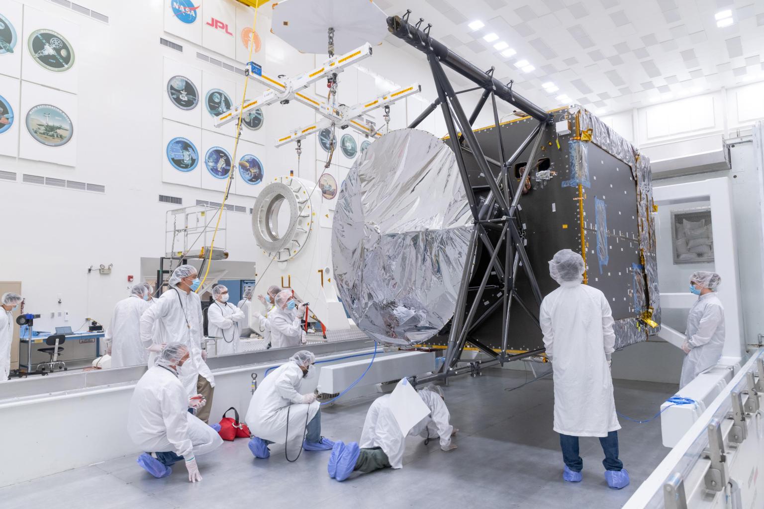 Several workers in white protective clothing and blue booties watch as engineers and technicians prepare to move the Solar Electric Propulsion (SEP) Chassis from its shipping container.