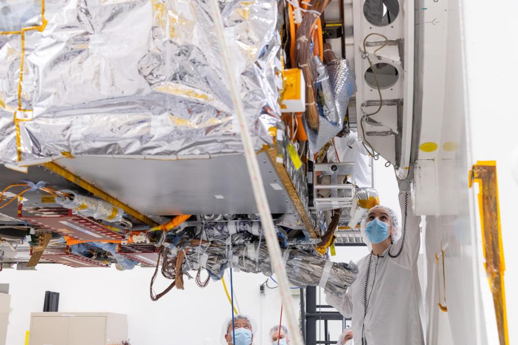 A worker in protective clothing looks up at the chassis of Psyche