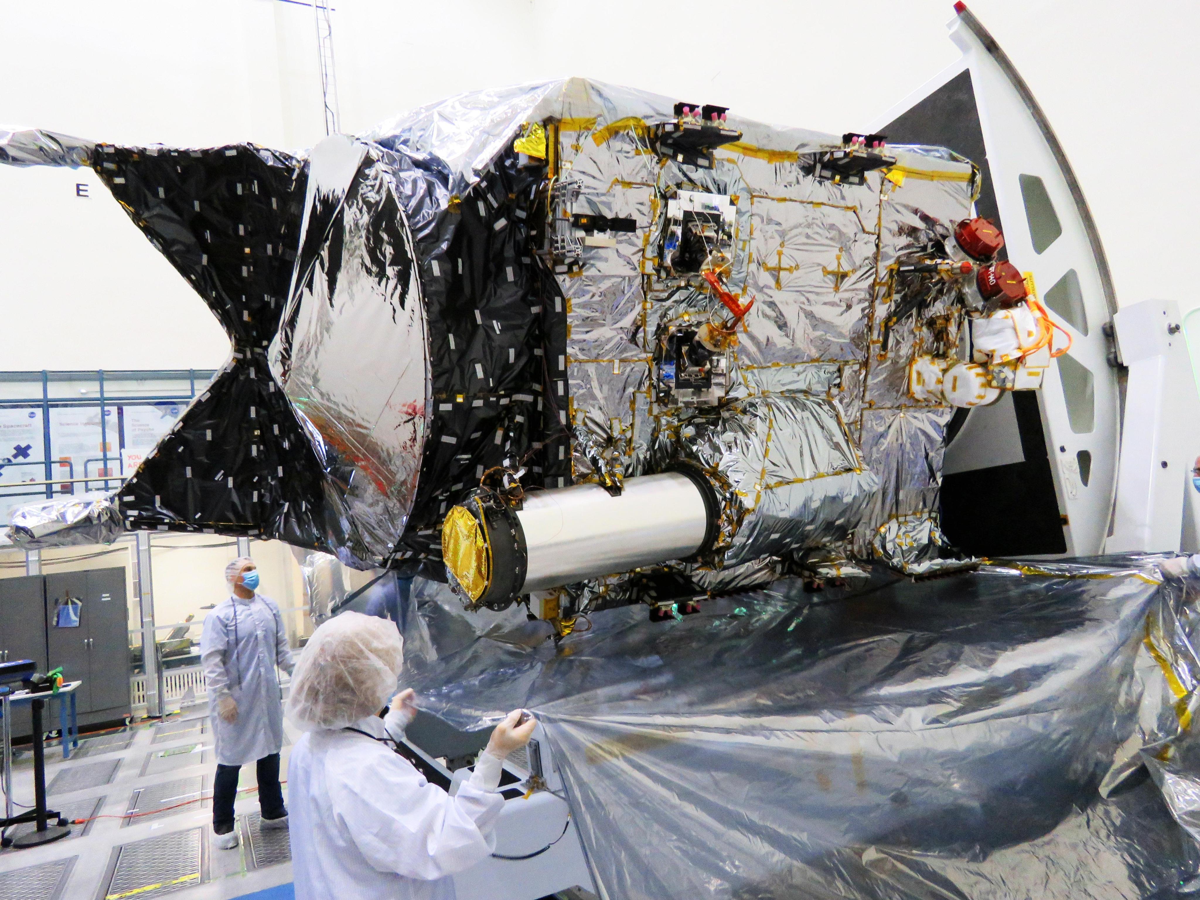 Workers stand near the Psyche spacecraft. One worker is holding the edges of a protective cover.