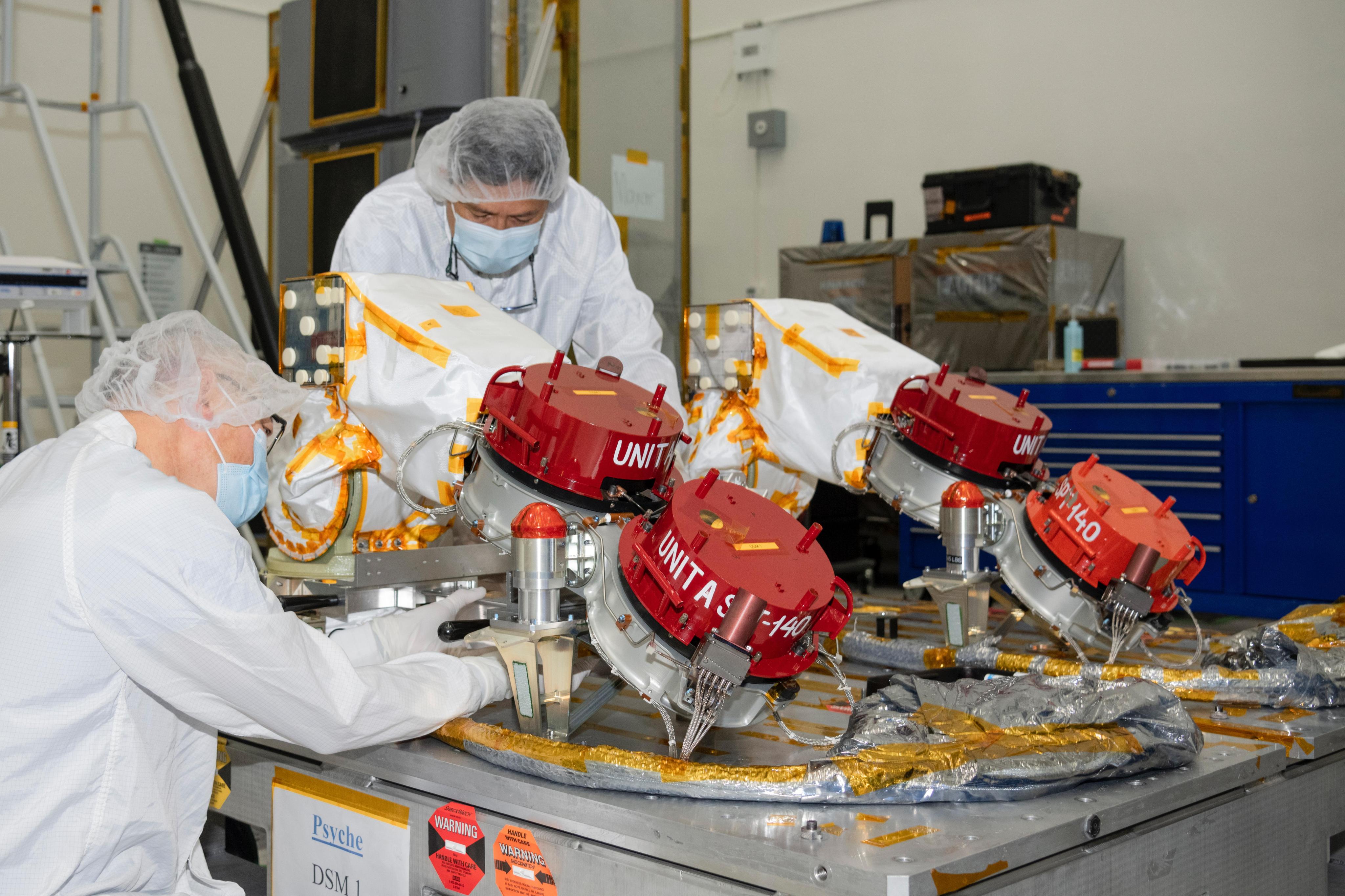 Two engineers in protective clothing work on part of the Psyche spacecraft. Red protective covers can be seen covering the four Hall thrusters.