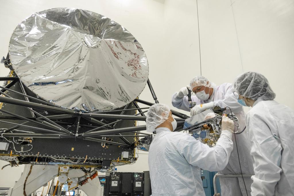 Workers in protective clothing hold an instrument and stand near a round, aluminum colored part of the spacecraft.