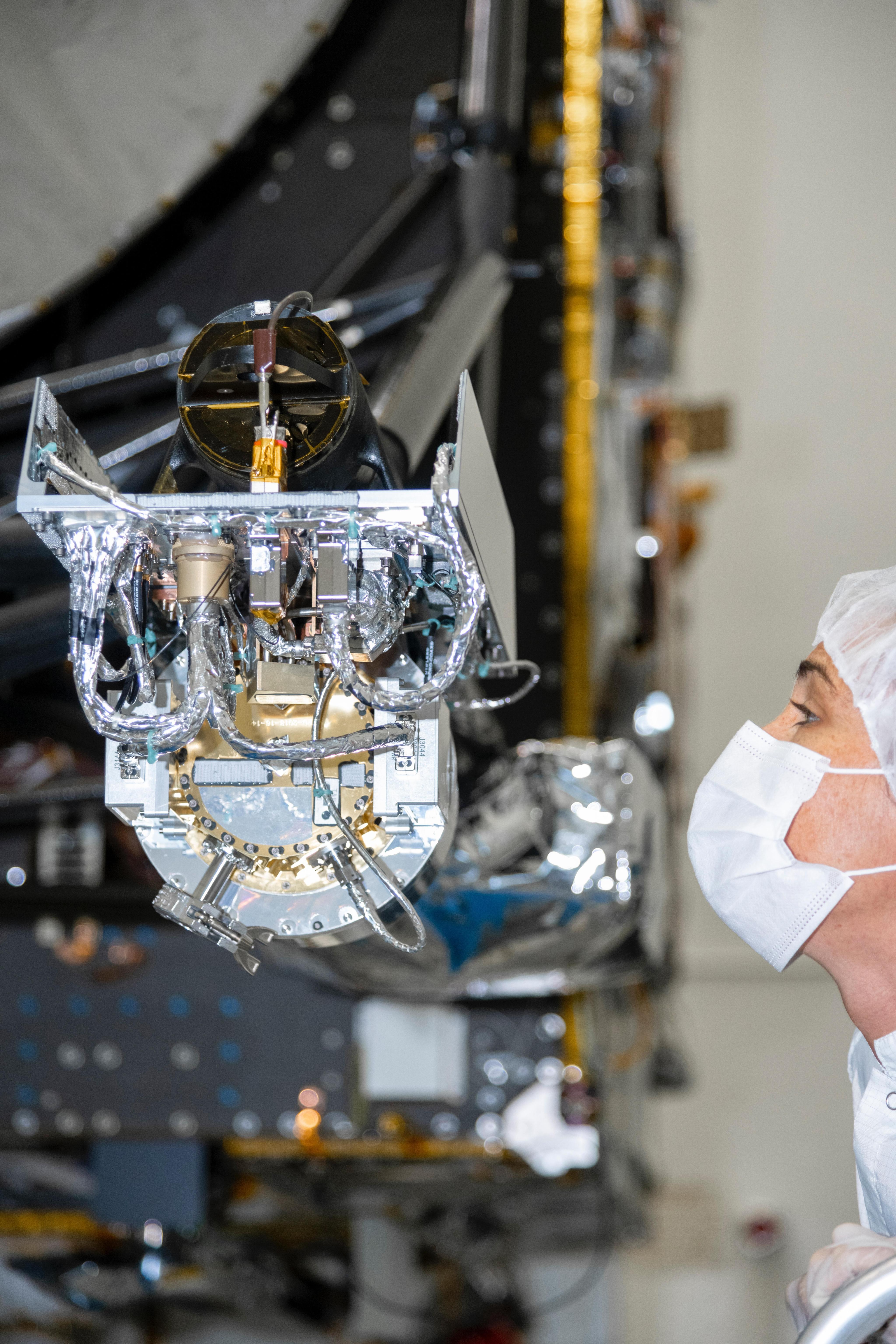 An engineer wearing a mask and head covering looks a spacecraft instrument with lots of wires visible.