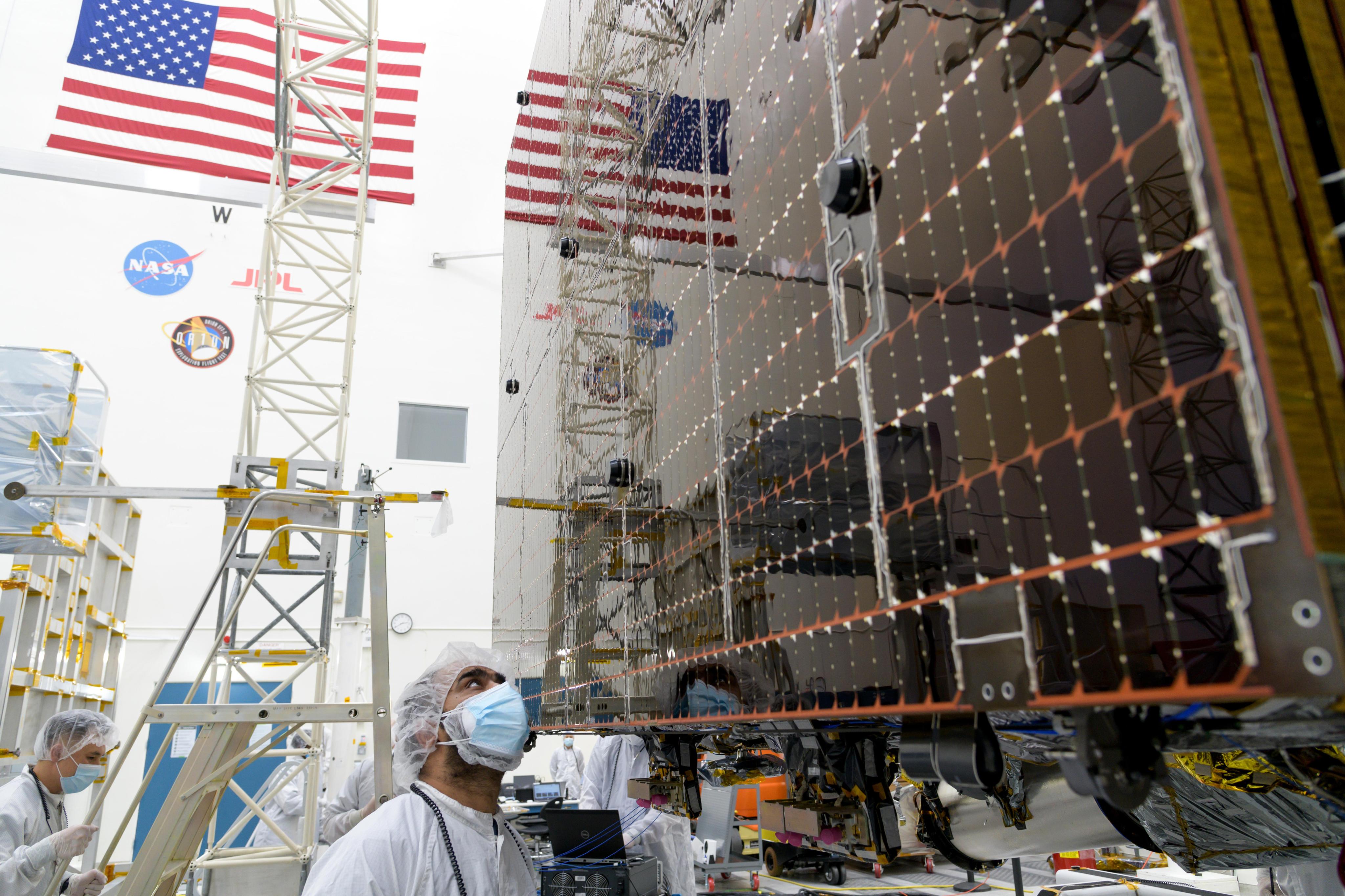 An engineer in white protective clothing, a head covering, and a blue face mask looks up at part of one of Psyche's solar panel.