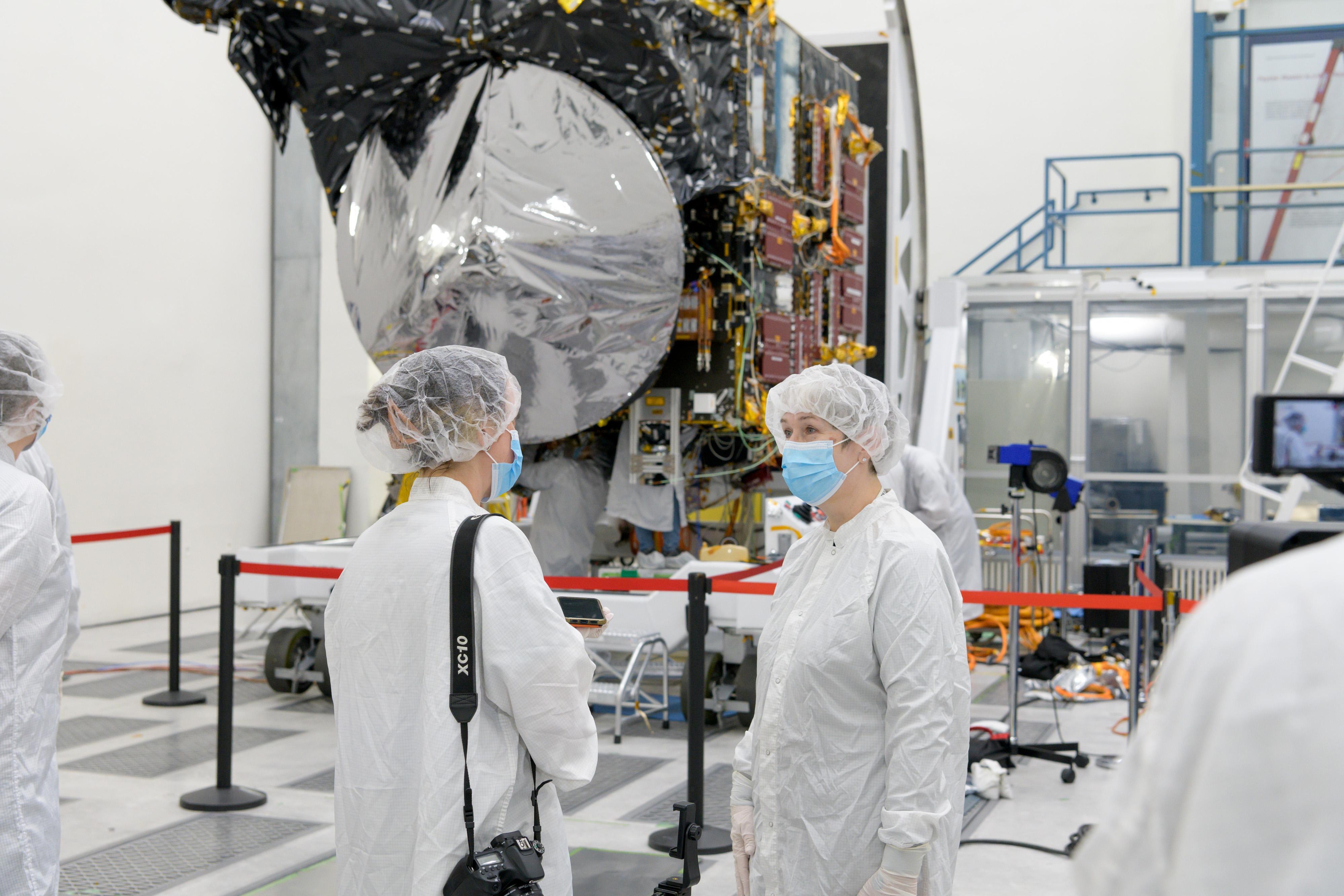 Two people dressed in white protective clothing and hair covers talk near the Psyche spacecraft.