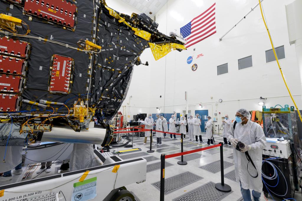 Members of the media stand behind the roped-off Psyche spacecraft. A worker is nearby with a piece of machinery.