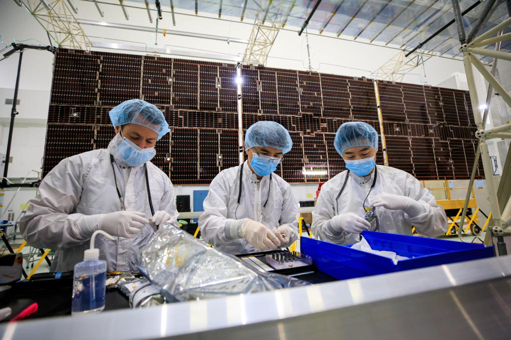 Three workers stand at a counter with parts and trays.