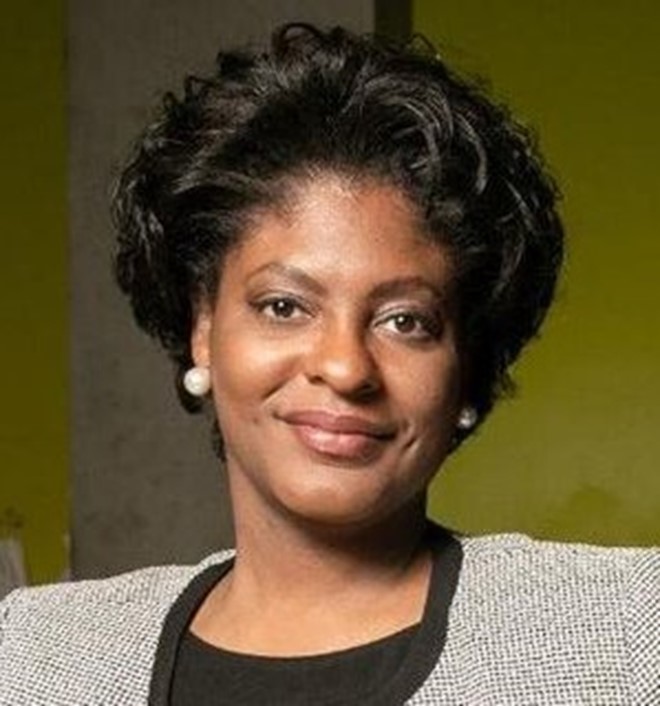 Portrait photo of a person with earrings and short hair wearing a patterned blazer over a dark shirt.