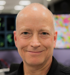 Portrait photo of a person with short hair wearing a collared shirt; a blurred multi-colored item is visible in the background.