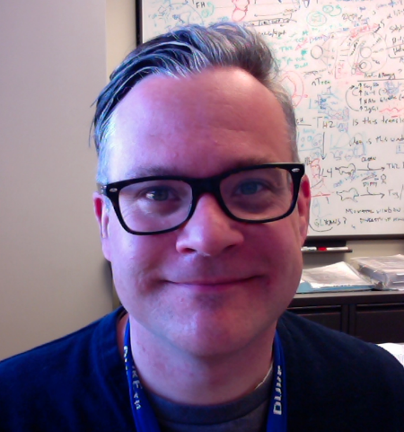 Portrait photo of a person with short hair wearing glasses and a lanyard; a whiteboard with symbols is visible in the background.