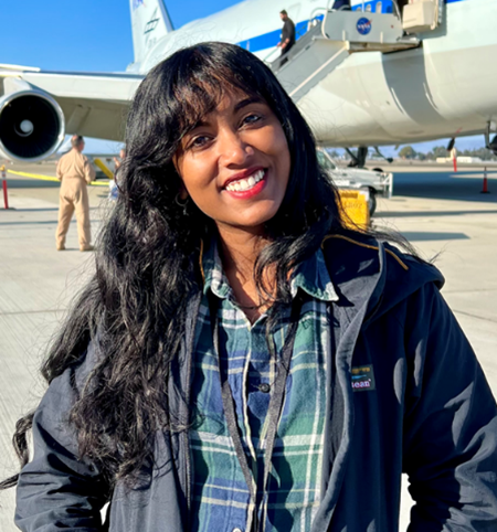 Portrait photo of a person with long hair wearing a plaid collared shirt and a jacket; an airplane is visible in the background.