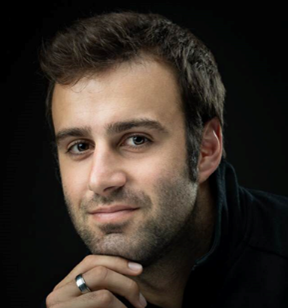 Portrait photo of a person with short hair wearing a dark shirt; a metal ring is visible on their hand touching their chin.