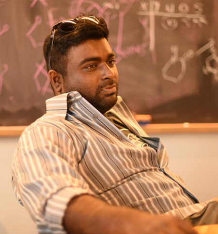 Portrait photo of a person with short hair and sunglasses on top of their head wearing a collared button-up striped shirt; a chalkboard with symbols is visible in the background.