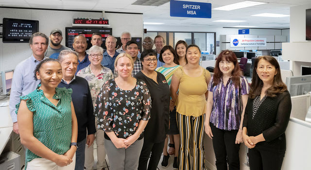 A group of 19 adults of various ages, smiling and dressed in casual business attire, stand together for a group photo in an office, amid desks and computer terminals. Signs hanging in the room read Spitzer MSA (for mission support area) and NASA-Jet Propulsion Laboratory-California Institute of Technology.